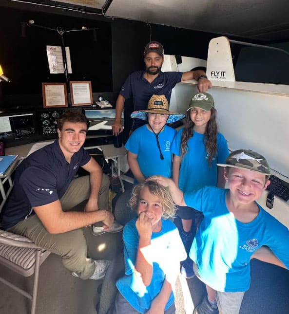 A group of students and a couple of adults stand in a room with a flight simulator